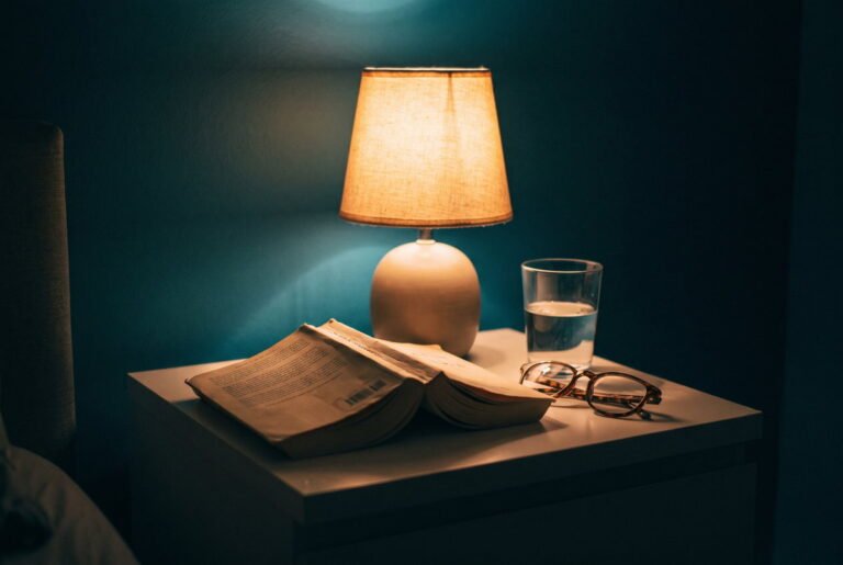 Una mesilla de noche con la lámpara encendida y un libro de bolsillo boca abajo, en una habitación a oscuras.